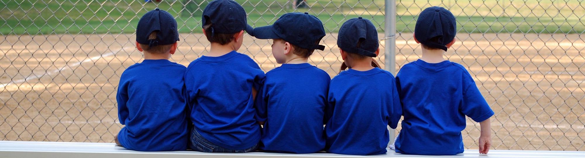 Rear view of a row of kids in blue baseball caps and T-shirts sitting on a bench at a ballfield