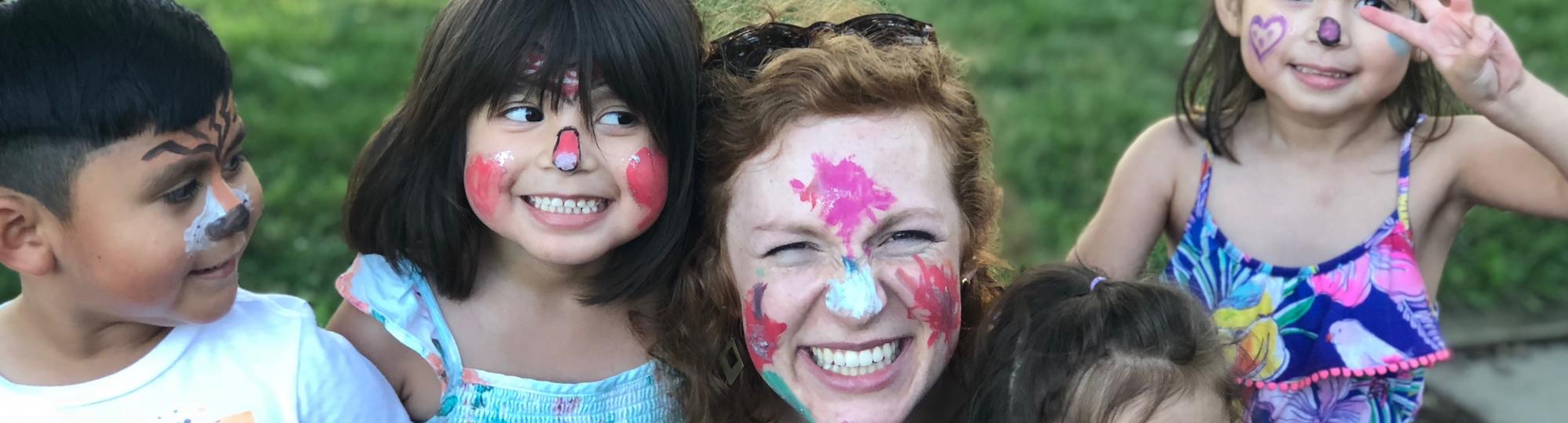 Young kids and a young woman outdoors on grass, wearing colorful face paint