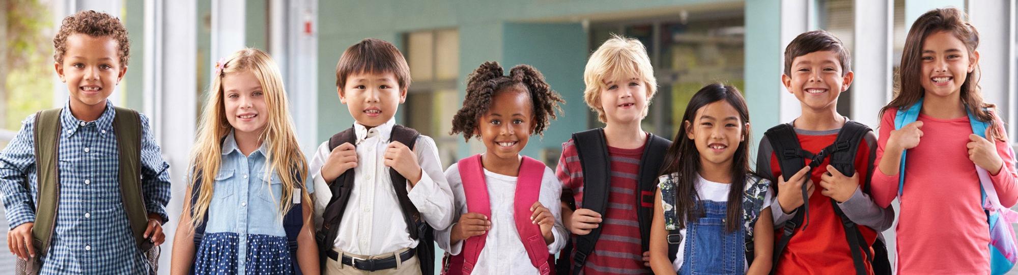 Smiling, ethnically diverse schoolchildren in colorful clothes and backpacks are lined up side by side