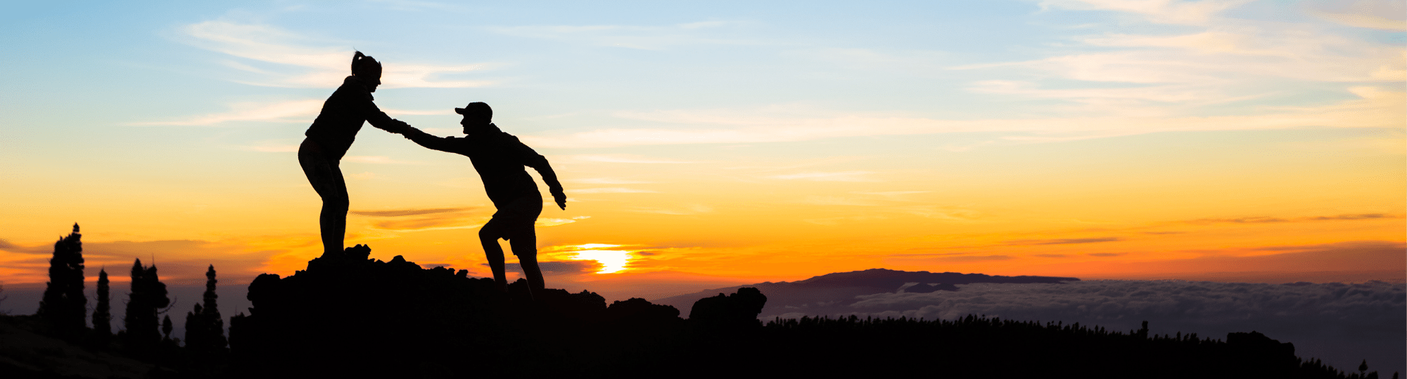 Silhouette of one person helping another to the top of a hill, with sunset behind them