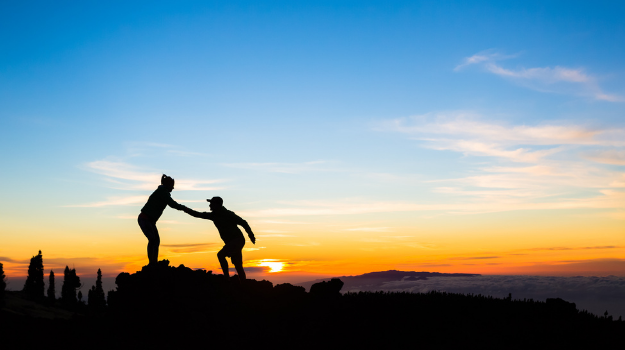 Silhouette of one person pulling another up to the top of a hill with sunset behind them