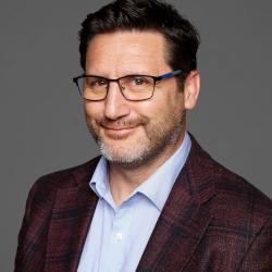 Nathan wears a brown suit jacket and glasses and looks at the camera